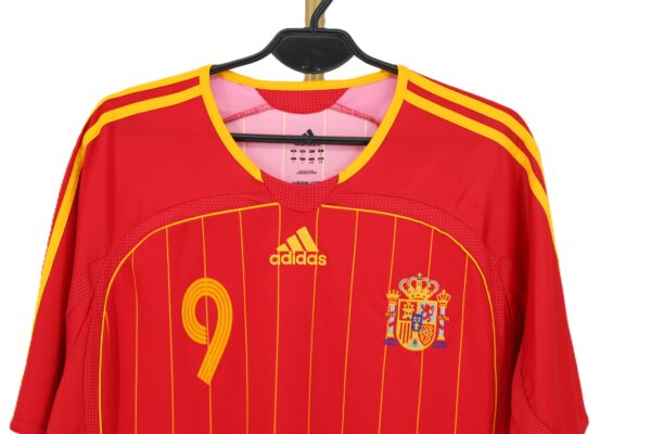 Zbliżenie na wyszywane godło Hiszpanii i żółte logo Adidas na czerwonej koszulce meczowej.